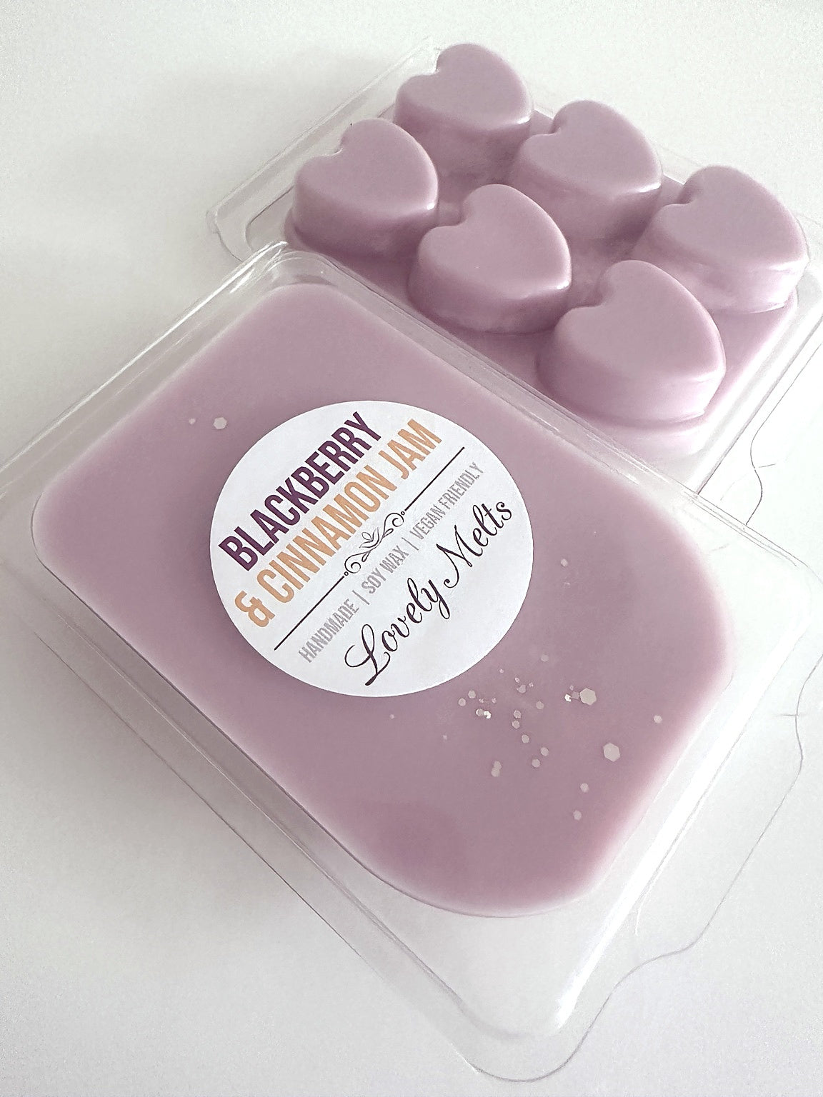 A clear plastic clamshell package of wax melts with a round white label reading 'Blackberry & Cinnamon Jam' and 'Lovely Melts,' shown alongside a back image of heart-shaped wax melts on a white background.