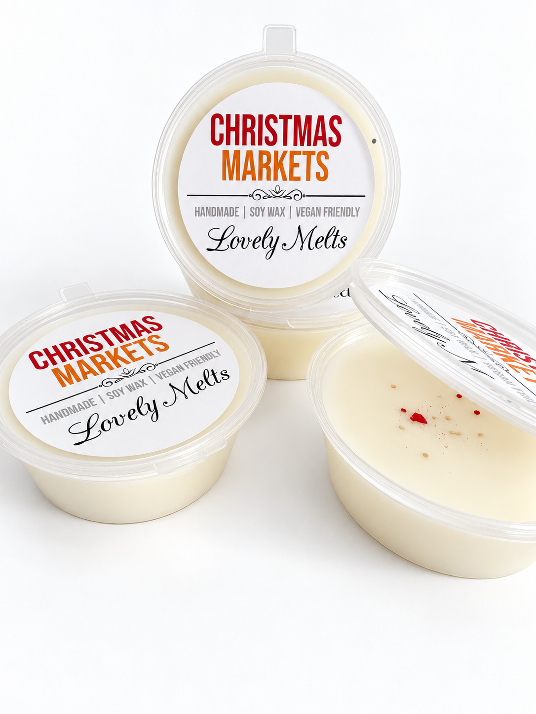 Three small round plastic containers of creamy off-white  soy wax melts with clear lids, arranged on a white background. Two lids show printed labels reading 'CHRISTMAS MARKETS' and 'Lovely Melts'; one container is open, showing the wax inside and a small red spice-like glitter on top.