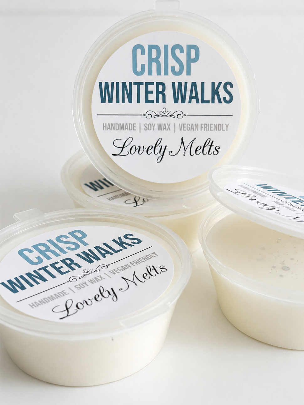 Four round plastic containers of soy wax melts with clear lids, showing the off-white wax inside. The lids feature a printed label for 'Crisp Winter Walks' by 'Lovely Melts' in blue and black text.