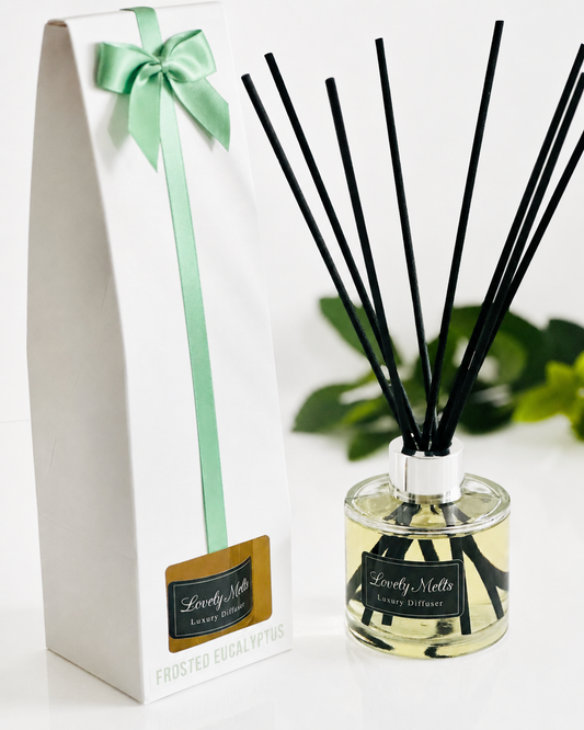 Reed diffuser with black sticks in a glass bottle, labeled 'Lovely Melts', next to its packaging with a green ribbon.