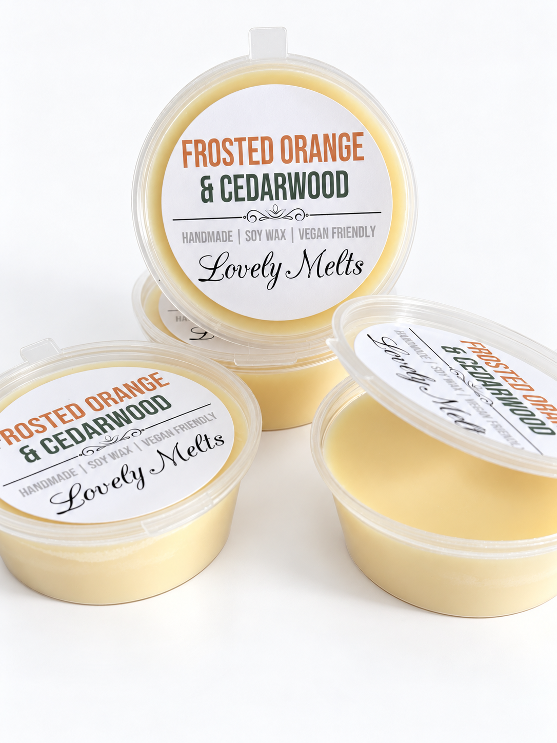Four small round plastic containers of soy wax melts on a white background, with one container open to show the pale orange wax inside. The labels read 'Frosted Orange & Cedarwood' and 'Lovely Melts'.