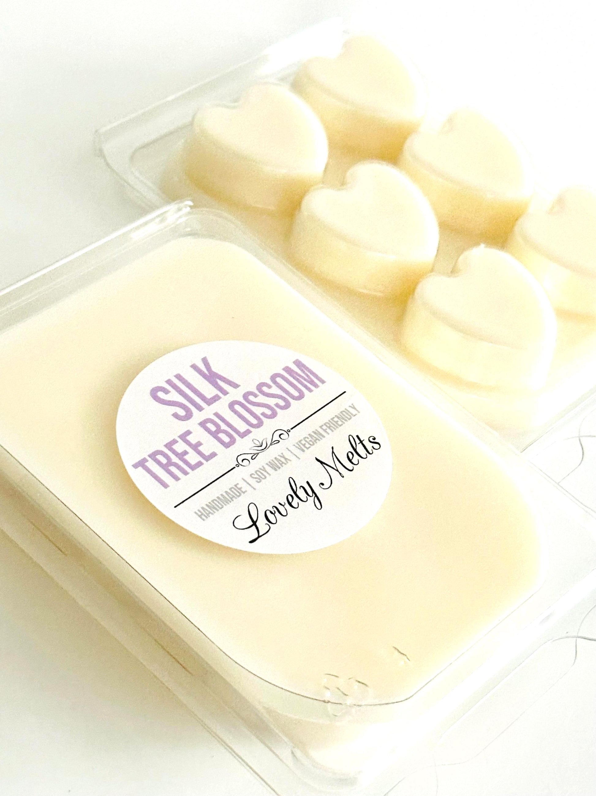 A clear plastic clamshell package of wax melts labeled 'SILK TREE BLOSSOM' by Lovely Melt's, with pale creamy wax melts visible inside and additional heart-shaped melts in the background.