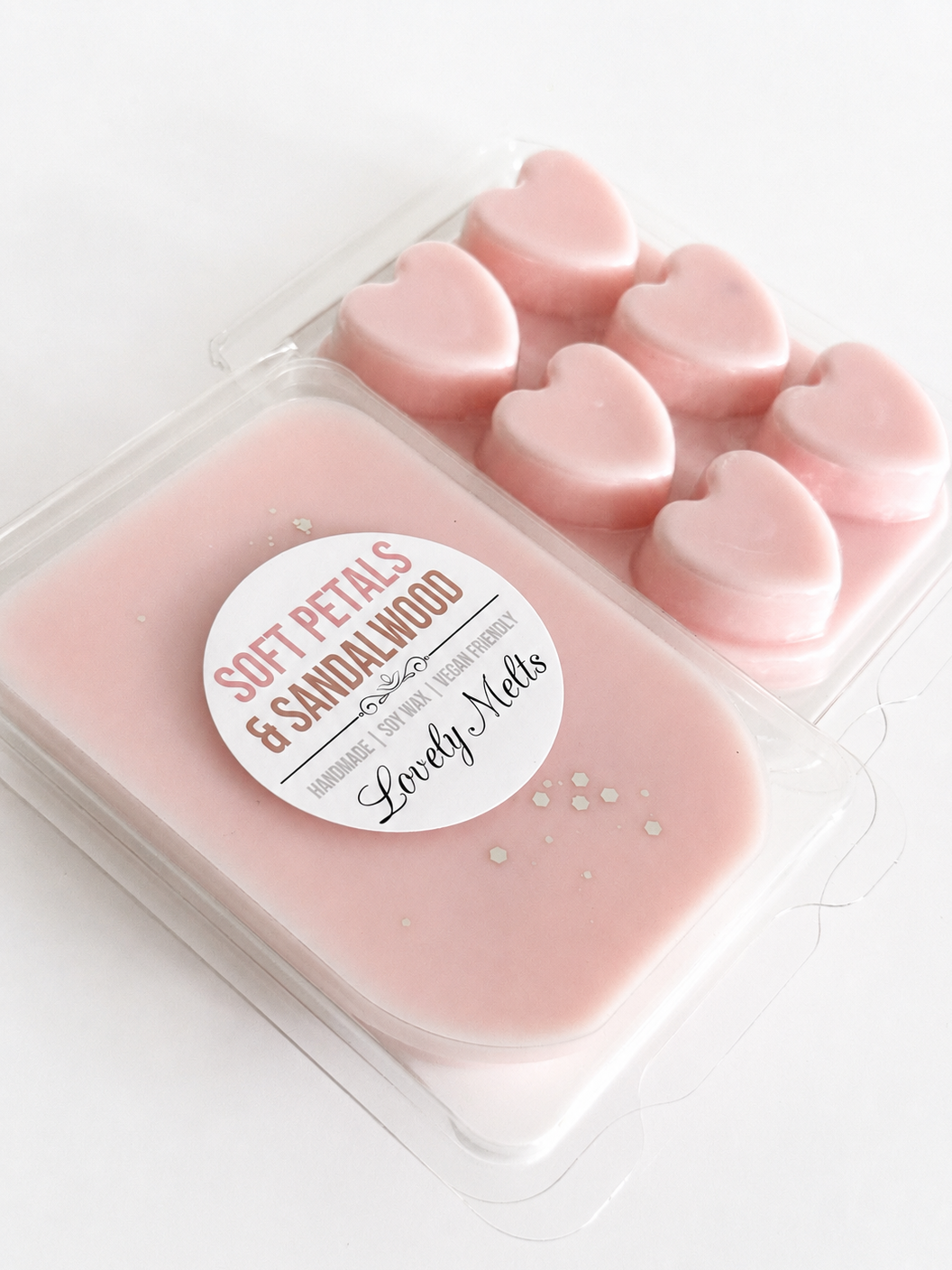 A clear plastic clamshell package of pink wax melts containing one large rectangular pink wax piece and six small heart-shaped pink wax pieces. A round white label reads 'SOFT PETALS & SANDALWOOD' and 'Lovely Melts'.