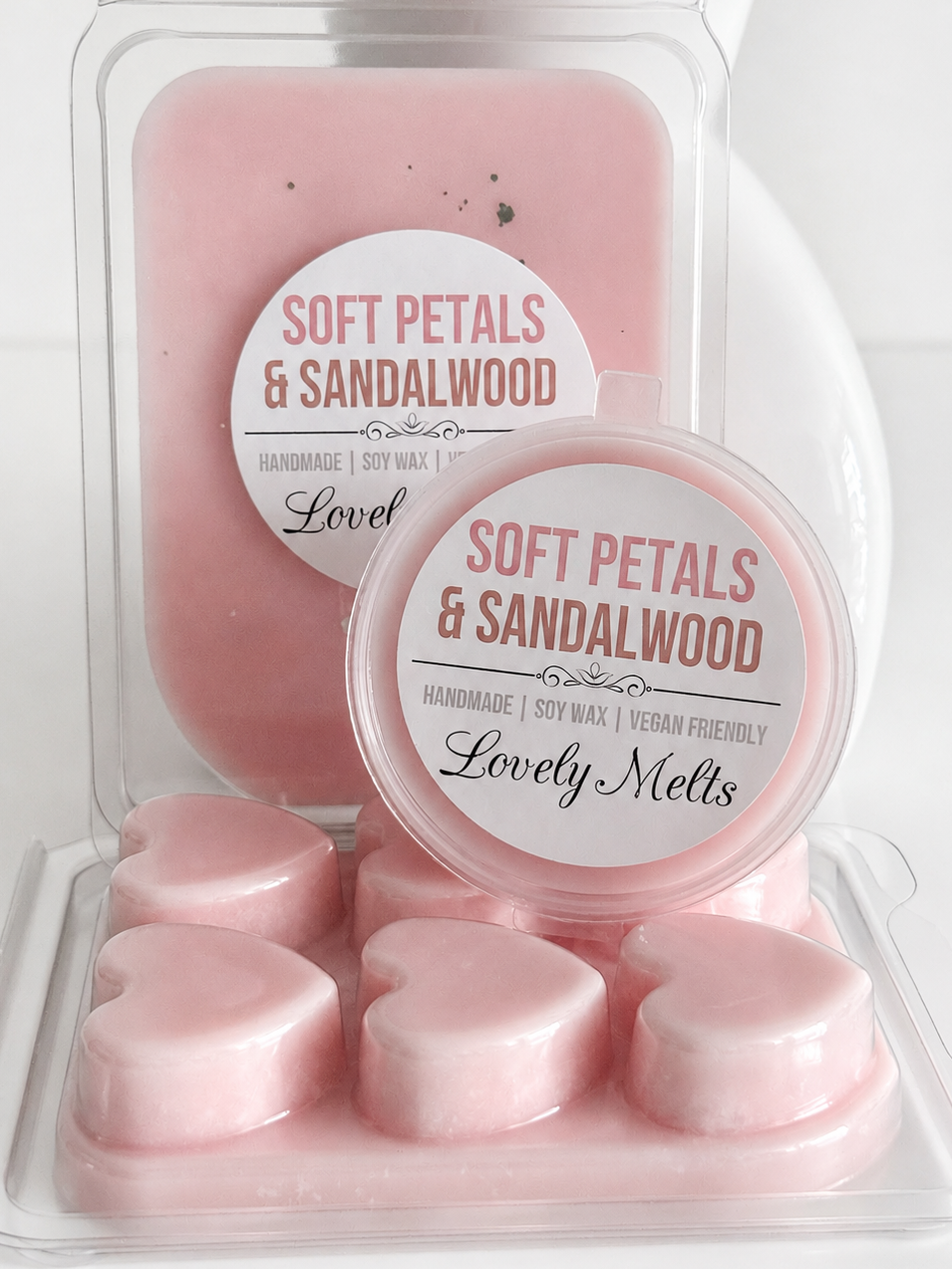 Pink soy wax melts and wax blocks in a clear plastic clamshell package, with round labels reading 'SOFT PETALS & SANDALWOOD' and 'Lovely Melts'.